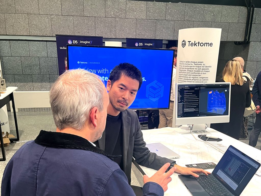 A Tektome team member talking with a visitor at the RELEASE [AEC] demo pod, with the review film on a large screen behind